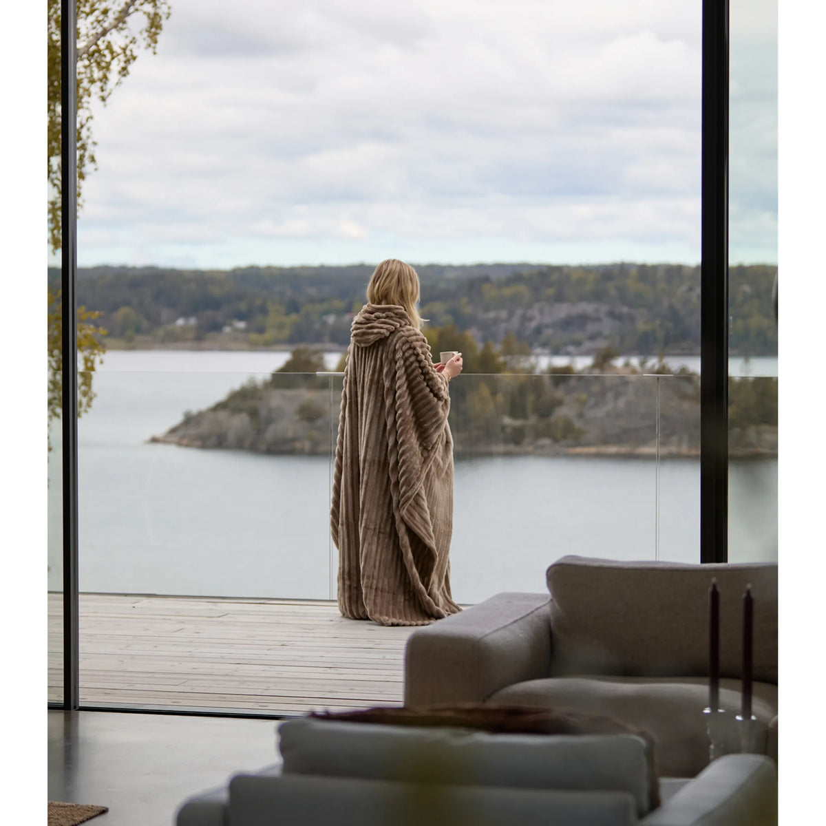 Henkilö, joka on pukeutunut Poncho Cozy -huppuponchoon, seisoo parvekkeella, josta avautuu näkymä järvelle.